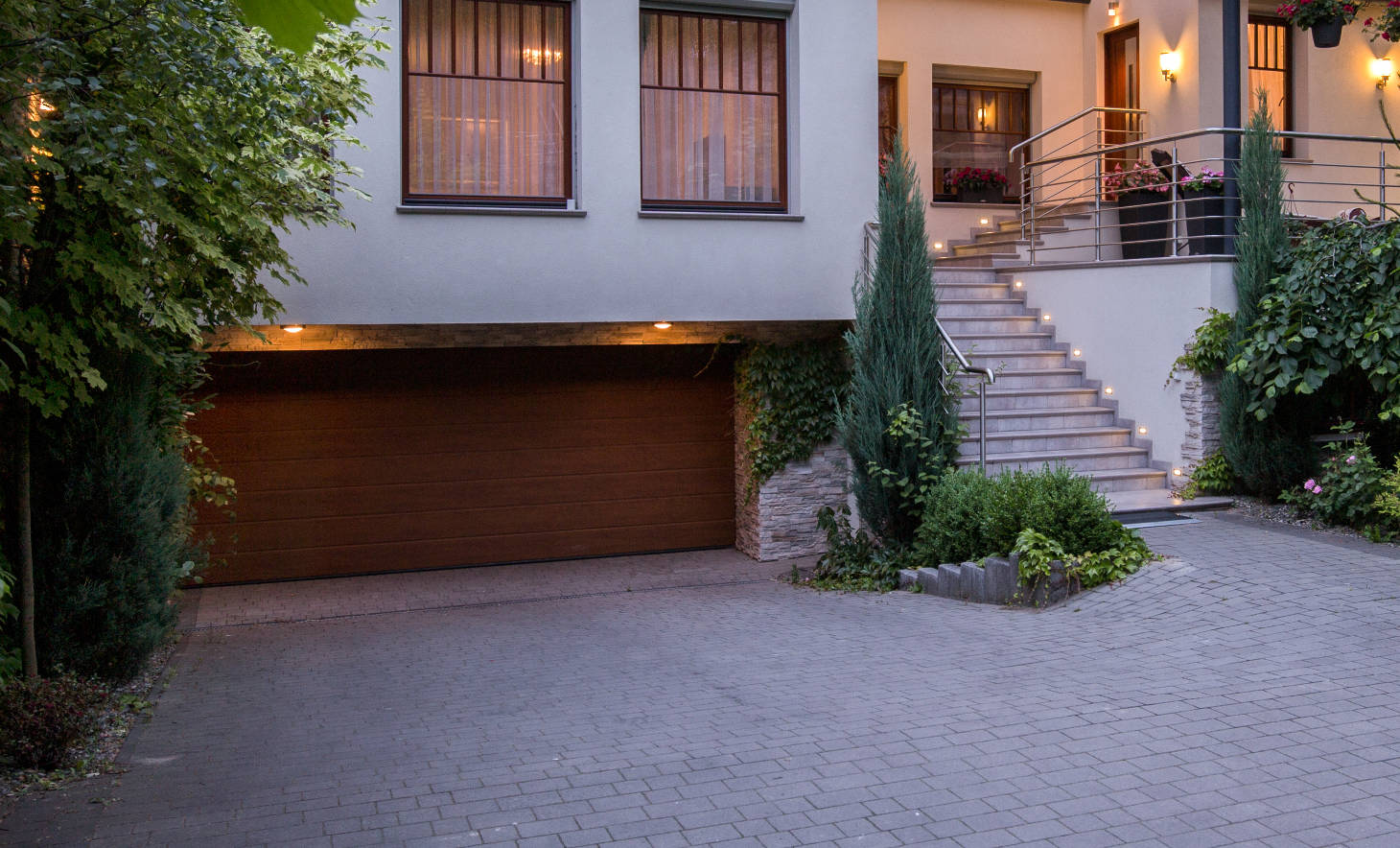 A modern home with a sleek design featuring a driveway paved with gray concrete pavers leading to a wooden garage door. The entryway includes illuminated steps with stainless steel railings, surrounded by neatly landscaped greenery, including shrubs and climbing vines, creating an elegant and welcoming appearance.