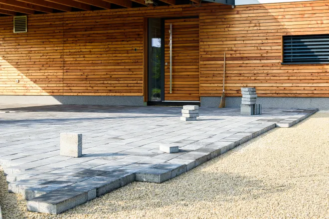 Interlocking stone driveway under construction in front of a modern wood-paneled home, with paving stones being carefully installed on a gravel base.